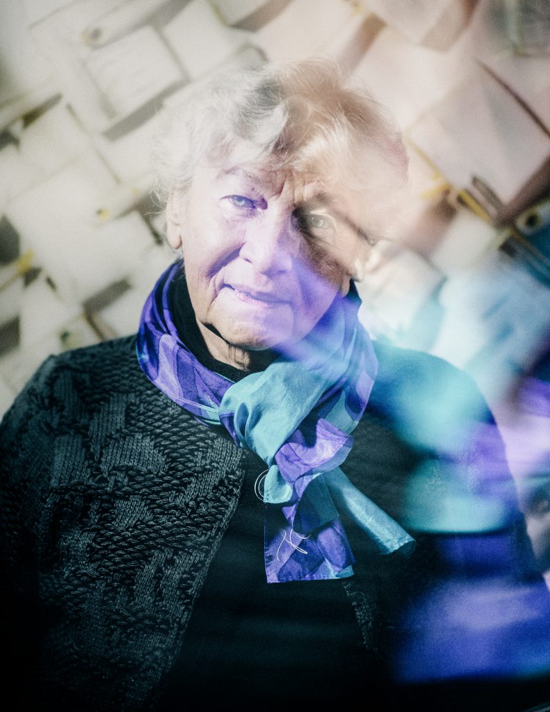 Colour photograph of Eliane Radigue at age 85, looking directly at the camera with her head tilted slightly to one side and the suggestion of a smile despite not being the biggest fan of having her photo taken. She wears a beautiful turquoise and magenta silk scarf which seems to be creating light reflections on the right hand side of the image.