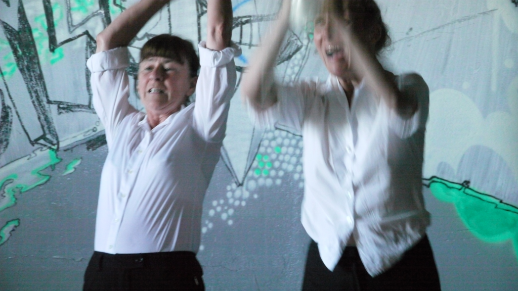 Photograph of Lovisa Johansson and Wenche Tankred in performance. They are wearing white shirts and their arms are raised in the midst of what appears to be some kind of ecstatic action