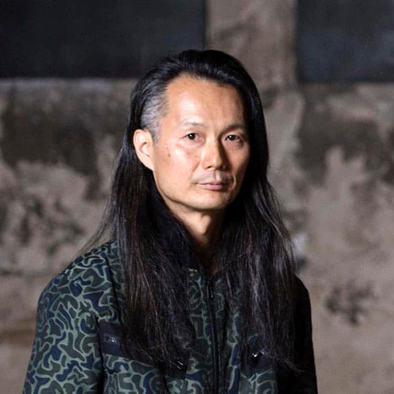 A person with long dark hair wearing a patterned jacket stands in front of a textured background.
