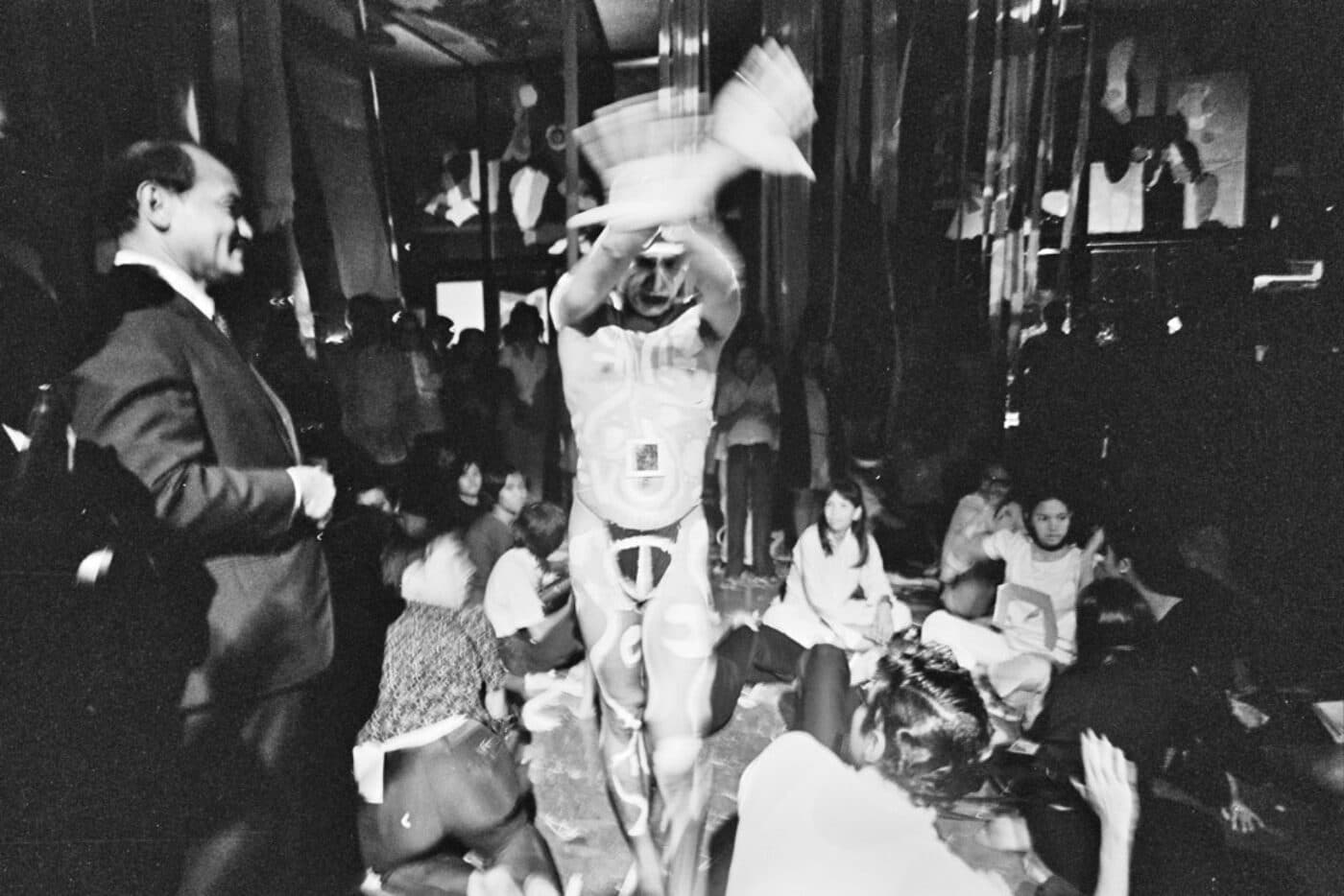 A person in motion is performing in front of a seated audience in a dimly lit room.