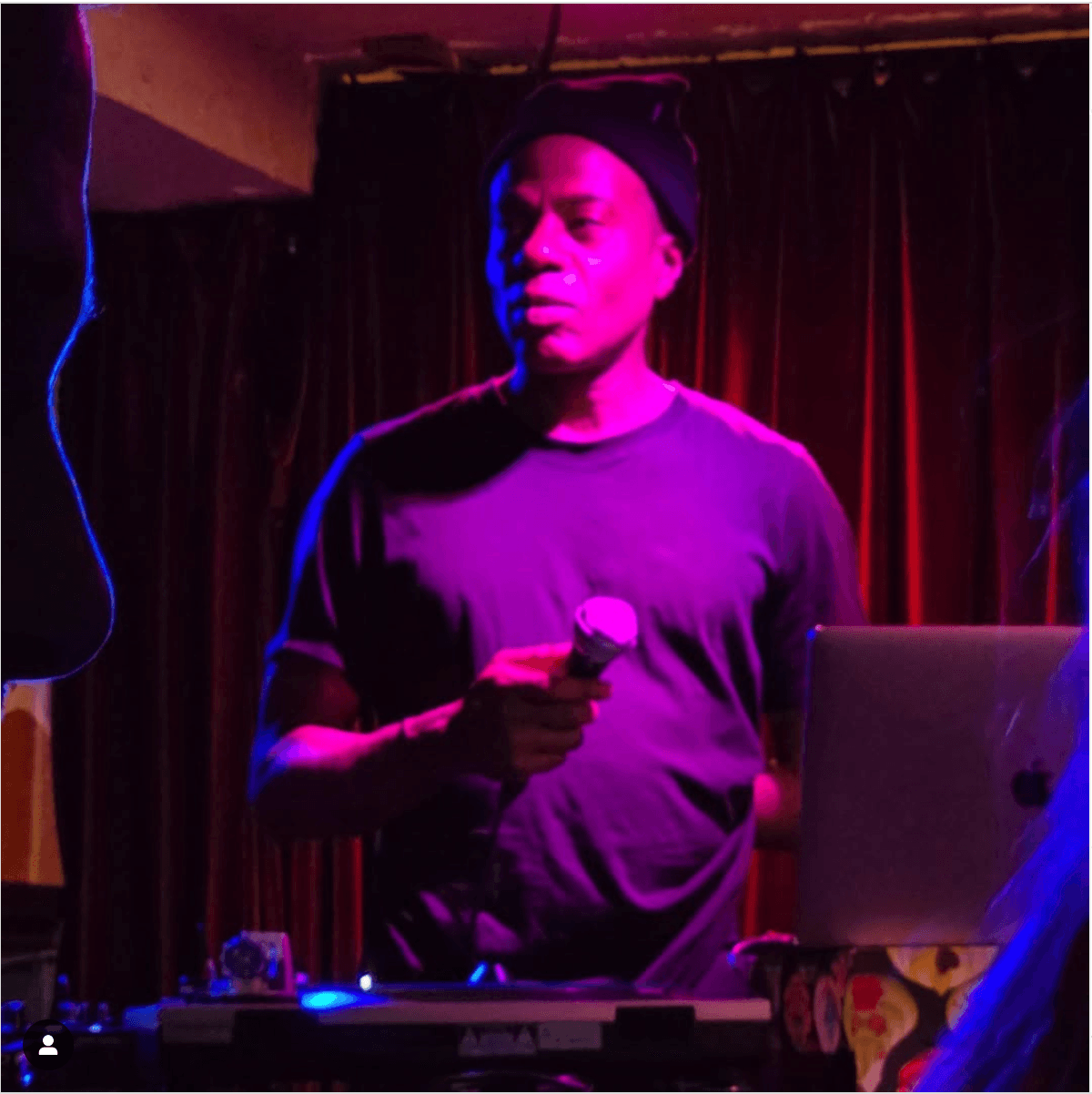 A person holding a microphone stands in front of DJ equipment, illuminated by purple and blue lighting with a curtain backdrop.