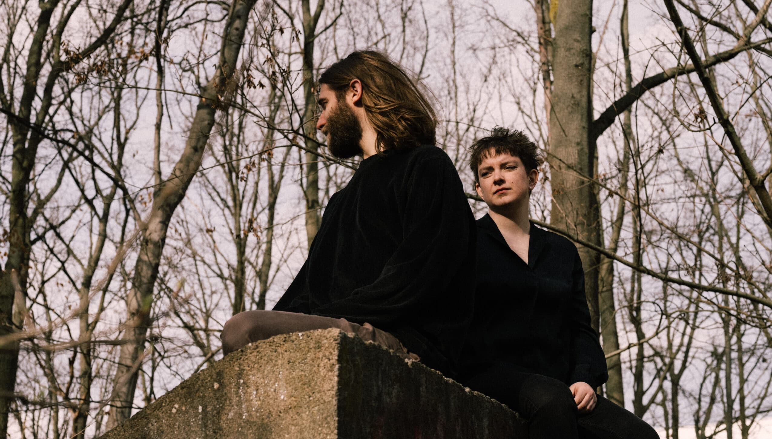 Two people are sitting on a concrete structure in a leafless forest, with one person facing away and the other looking forward.