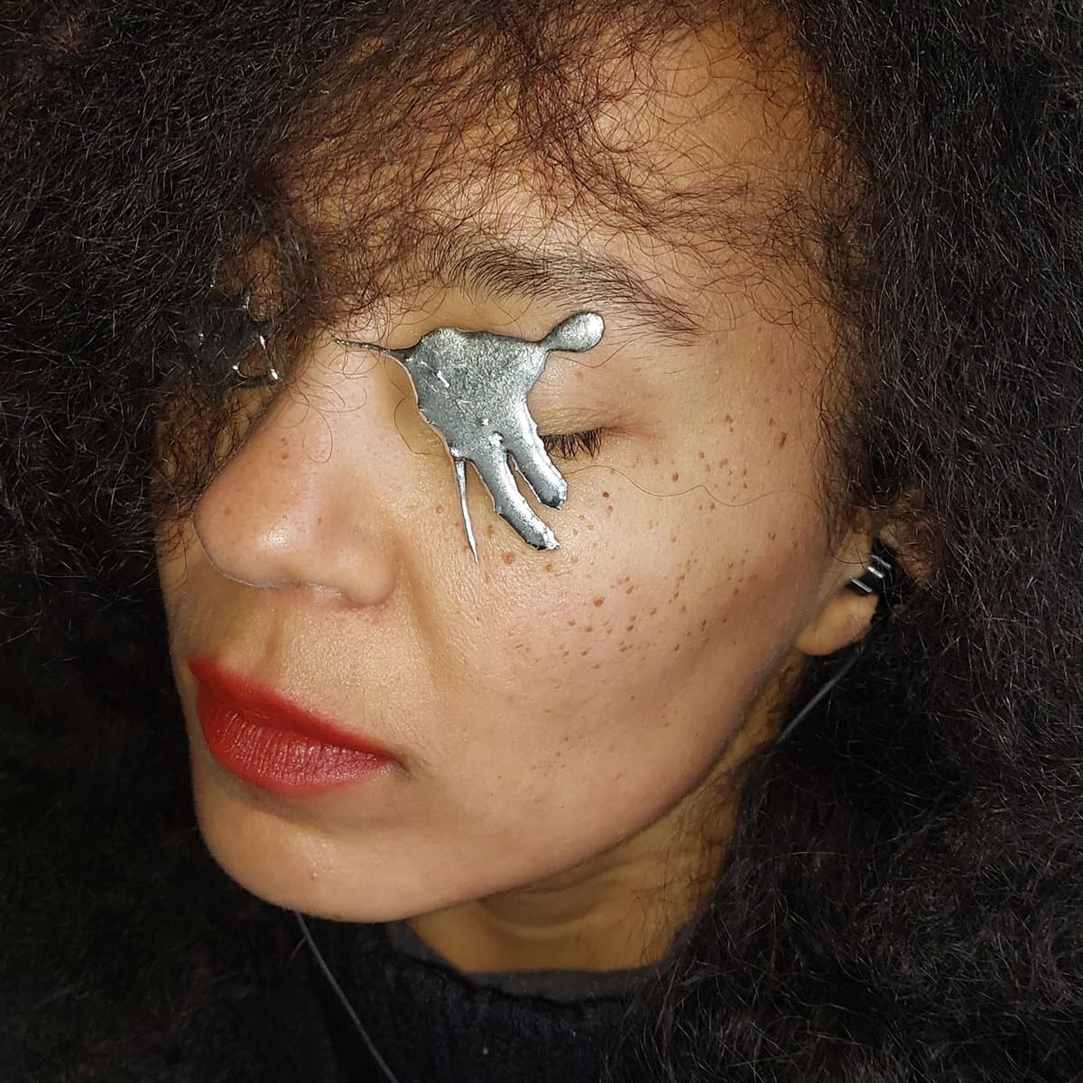 A person with curly hair and red lipstick has a metallic, hand-shaped object placed over their closed eye.